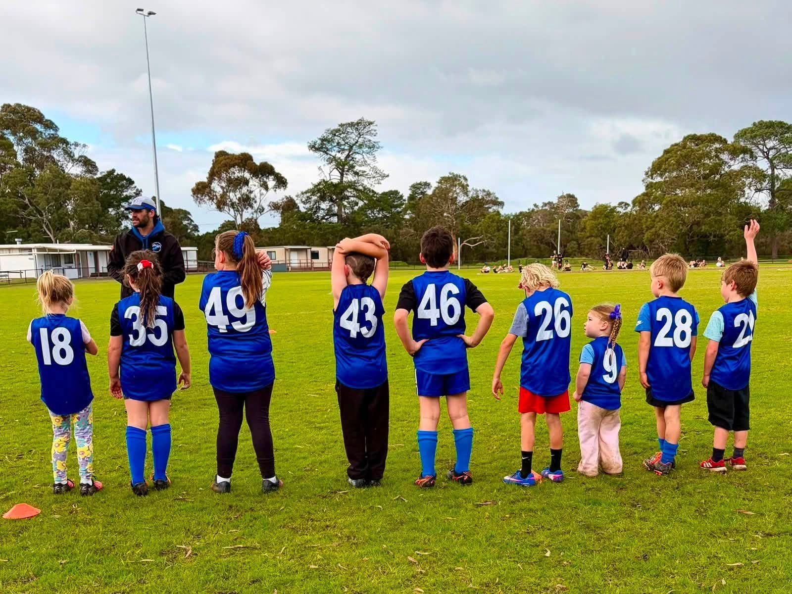 A row of Hastings juniors in blue guernseys stand side by side with their backs to the camera, listening to their coach at training.