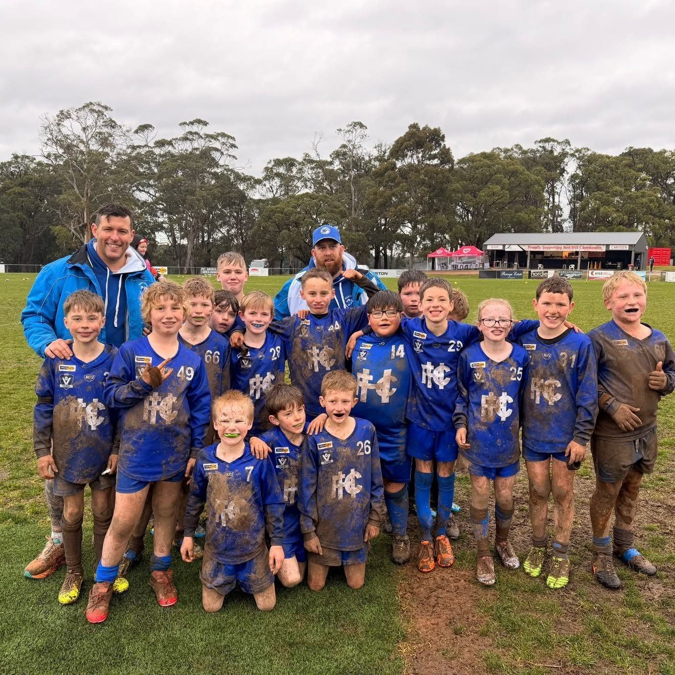 A muddy, smiling Hastings junior team lines up for a photo with their coaches after a wet game.